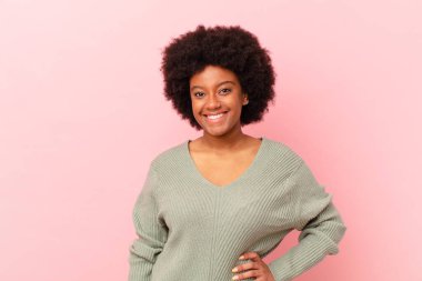 Afro siyah kadın mutlu bir şekilde gülümsüyor, eli kalçasında ve kendinden emin, olumlu, gururlu ve arkadaş canlısı.