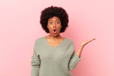 Afro siyahi kadın şaşırmış ve şok olmuş görünüyor, çenesi düşmüş, elinde açık bir elle bir nesne tutuyor.