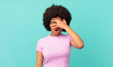 Afro siyah kadın şok olmuş, korkmuş ya da korkmuş görünüyor. Yüzü elle kaplanmış ve parmaklarının arasından gizlice bakıyor.