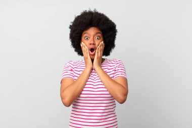 Afro siyah kadın şok olmuş ve korkmuş hissediyor, ağzı açık ve elleri yanaklarında korkmuş görünüyor.