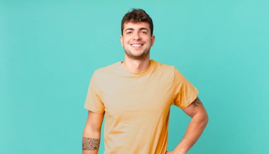 Yakışıklı adam mutlu bir şekilde gülümsüyor, eli kalçasında ve kendinden emin, olumlu, gururlu ve arkadaş canlısı.