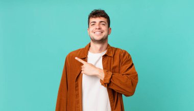 Yakışıklı adam neşeyle gülümsüyor, mutlu hissediyor ve yan ve yukarı doğru işaret ediyor, fotokopi uzayındaki nesneleri gösteriyor.