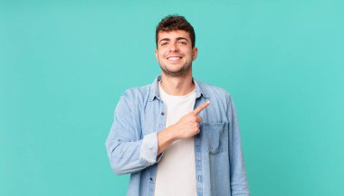 Yakışıklı adam neşeyle gülümsüyor, mutlu hissediyor ve yan ve yukarı doğru işaret ediyor, fotokopi uzayındaki nesneleri gösteriyor.