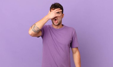 Yakışıklı adam şok olmuş, korkmuş ya da korkmuş görünüyor. Yüzü elle kaplanmış ve parmaklarının arasından gizlice bakıyor.
