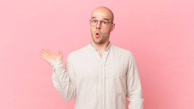 Kel adam şaşırmış ve şoka uğramış görünüyor, çenesi düşmüş ve eli açık bir nesneyi tutuyor.