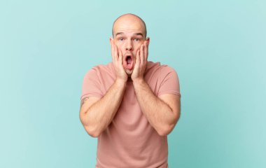 Kel adam şok olmuş ve korkmuş hissediyor, ağzı açık ve elleri yanaklarında korkmuş görünüyor.