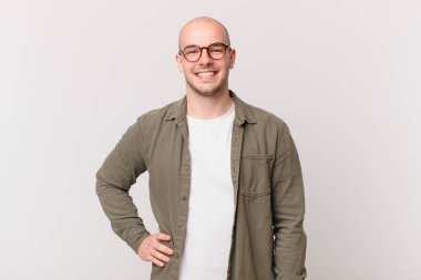 Kel adam mutlu bir şekilde gülümsüyor, eli kalçasında ve kendinden emin, olumlu, gururlu ve arkadaş canlısı.