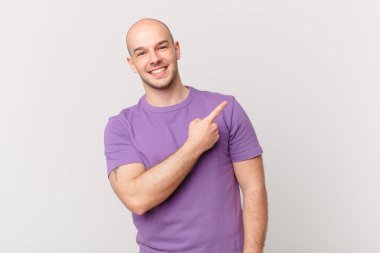 Kel adam neşeyle gülümsüyor, mutlu hissediyor ve kenarı ve yukarıyı işaret ediyor, fotokopi alanında nesneler gösteriyor.