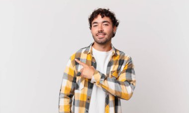 Hispanik yakışıklı adam neşeyle gülümsüyor, mutlu hissediyor ve kenarı ve yukarıyı işaret ediyor, fotokopi uzayında nesne gösteriyor