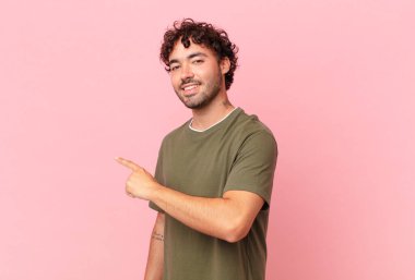 Hispanik yakışıklı adam neşeyle gülümsüyor, mutlu hissediyor ve kenarı ve yukarıyı işaret ediyor, fotokopi uzayında nesne gösteriyor