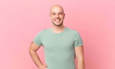 Kel adam mutlu bir şekilde gülümsüyor, eli kalçasında ve kendinden emin, olumlu, gururlu ve arkadaş canlısı.