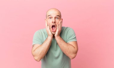 Kel adam şok olmuş ve korkmuş hissediyor, ağzı açık ve elleri yanaklarında korkmuş görünüyor.