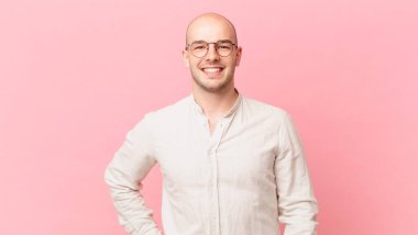 Kel adam mutlu bir şekilde gülümsüyor, eli kalçasında ve kendinden emin, olumlu, gururlu ve arkadaş canlısı.