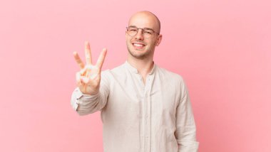 Kel adam gülümsüyor ve arkadaş canlısı görünüyor, üç ya da üç numarayı gösteriyor, eli önde, geriye doğru sayıyor.