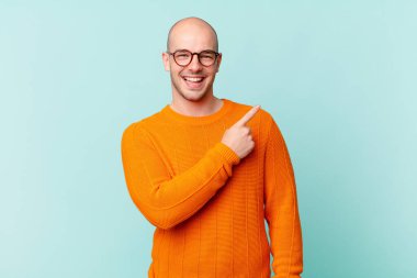 Kel adam neşeyle gülümsüyor, mutlu hissediyor ve kenarı ve yukarıyı işaret ediyor, fotokopi alanında nesneler gösteriyor.