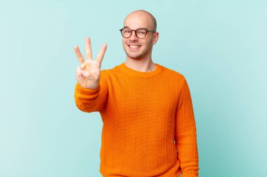 Kel adam gülümsüyor ve arkadaş canlısı görünüyor, üç ya da üç numarayı gösteriyor, eli önde, geriye doğru sayıyor.