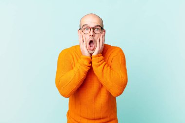 Kel adam şok olmuş ve korkmuş hissediyor, ağzı açık ve elleri yanaklarında korkmuş görünüyor.