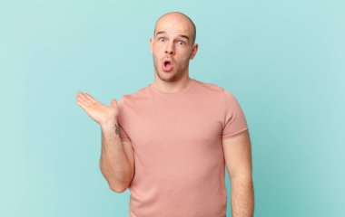 Kel adam şaşırmış ve şoka uğramış görünüyor, çenesi düşmüş ve eli açık bir nesneyi tutuyor.