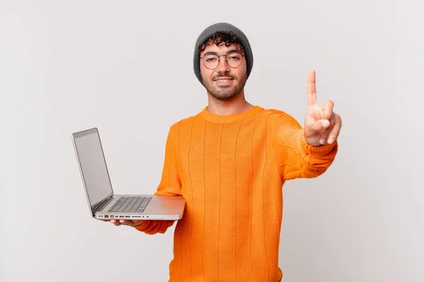 コンピュータを持つオタクの男は笑顔で友好的に見えます前方に手で1番または1番を示します — ストック写真