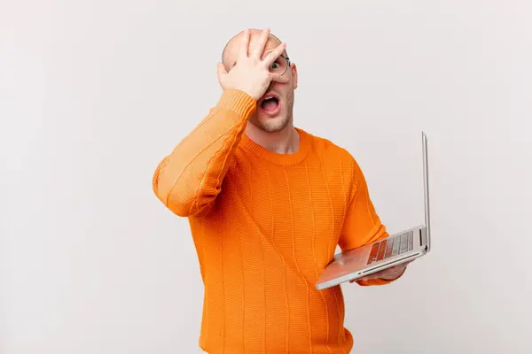 衝撃を受け 怖がったり恐れたり 手で顔を覆ったり 指の間を覗いたりしているコンピュータを持つハゲ男 — ストック写真