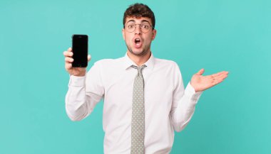 Akıllı telefonlu bir iş, şaşırmış ve şok olmuş görünüyor, çenesi düşmüş, bir objeyi elinde açık bir şekilde tutuyor.