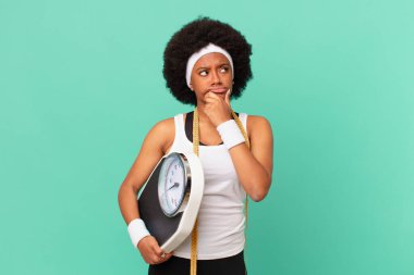 Afro kadın düşünüyor, kuşkulu ve şaşkın hissediyor, farklı seçeneklerle, hangi diyet konseptini seçeceğini merak ediyor.