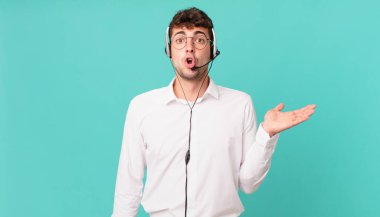 Tele-pazarlamacı şaşırmış ve şok olmuş görünüyor, çenesi düşmüş, bir objeyi elinde açık bir elle tutuyor.