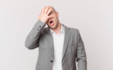Şok olmuş, korkmuş ya da korkmuş görünen kel iş adamı elleriyle yüzünü kapatıyor ve parmaklarının arasından gözetliyor.