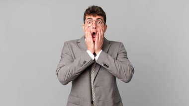 İş adamı şok olmuş ve korkmuş hissediyor, ağzı açık ve elleri yanaklarında korkmuş görünüyor.