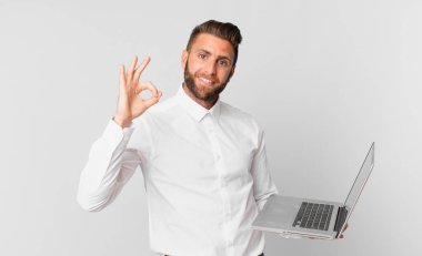 Genç ve yakışıklı bir adam kendini mutlu hissediyor. Onayını veriyor. İyi bir jest yapıyor ve elinde dizüstü bilgisayar tutuyor.