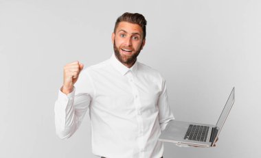 Genç yakışıklı adam şok olmuş hissediyor, gülüyor, başarıyı kutluyor ve elinde bir dizüstü bilgisayar tutuyor.