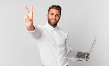 Genç yakışıklı adam gülümsüyor ve mutlu görünüyor. Zafer ya da barış işareti yapıyor ve elinde bir dizüstü bilgisayar tutuyor.