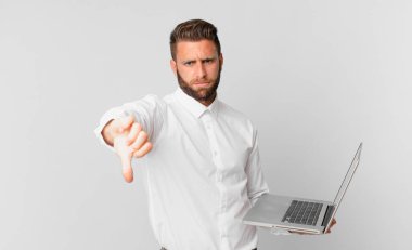 Genç ve yakışıklı adam huysuz hissediyor, baş parmaklarını gösteriyor ve elinde bir dizüstü bilgisayar tutuyor.