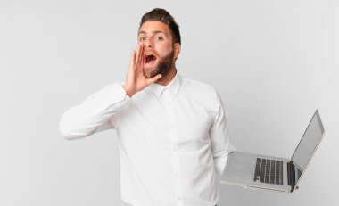 Genç yakışıklı adam mutlu hissediyor, elleri ağızının yanında ve elinde dizüstü bilgisayarla bağırıyor.