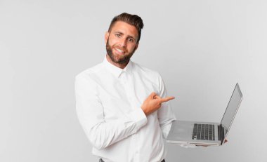 Genç ve yakışıklı bir adam neşeyle gülümsüyor, mutlu hissediyor ve yan tarafı işaret ediyor ve elinde bir dizüstü bilgisayar tutuyor.