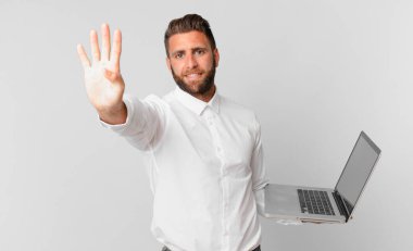 Genç yakışıklı adam gülümsüyor ve arkadaş canlısı görünüyor, dört numarayı gösteriyor ve elinde bir dizüstü bilgisayar var.