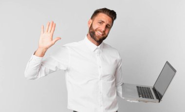 Genç yakışıklı adam mutlu bir şekilde gülümsüyor, el sallıyor, seni selamlıyor ve dizüstü bilgisayarını tutuyor.