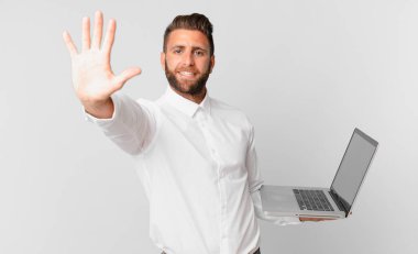Genç yakışıklı adam gülümsüyor ve arkadaş canlısı görünüyor, beş numarayı gösteriyor ve elinde bir dizüstü bilgisayar var.