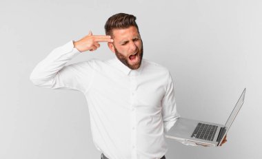 Genç yakışıklı adam mutsuz ve stresli görünüyor, intihar jesti silah işareti yapıyor ve elinde bir dizüstü bilgisayar tutuyor.