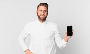 Genç yakışıklı adam mutlu bir şekilde gülümsüyor, eli kalçasında, kendinden emin ve elinde bir hücreyle.