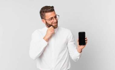 Mutlu ve kendinden emin bir ifadeyle gülümseyen genç yakışıklı bir adam. Eli çenesinde ve elinde hücresiyle.