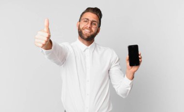 Genç yakışıklı adam gururlu, olumlu gülümsüyor, parmakları havada ve elinde bir hücreyle.