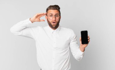 Genç yakışıklı adam mutlu görünüyor, şaşırmış ve şaşırmış ve elinde bir hücre tutuyor.