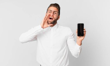 Genç yakışıklı adam mutlu hissediyor, elleri ağızının yanında ve elinde bir hücreyle bağırıyor.