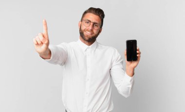 Genç yakışıklı adam gülümsüyor ve arkadaş canlısı görünüyor. Bir numarayı gösteriyor ve elinde bir hücre tutuyor.