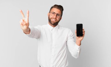 Genç yakışıklı adam gülümsüyor ve arkadaş canlısı görünüyor. İki numarayı gösteriyor ve elinde bir hücre tutuyor.