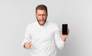 Genç yakışıklı adam agresif bir şekilde bağırıyor, kızgın görünüyor ve elinde bir hücre tutuyor.