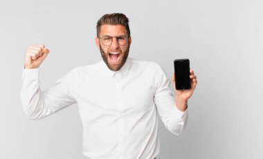 young handsome man shouting aggressively with an angry expression and holding a cell