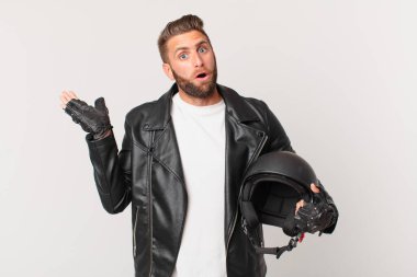young handsome man looking surprised and shocked, with jaw dropped holding an object. motorbike helmet concept