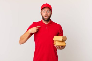 young handsome man looking shocked and surprised with mouth wide open, pointing to self burger delivering concept